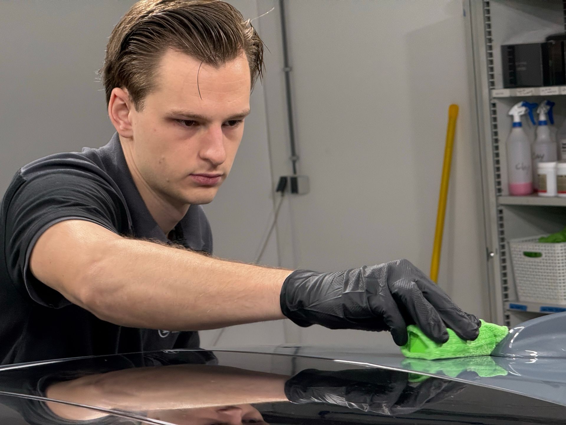 Person wearing black gloves cleaning a car surface with a green microfiber cloth in a workshop.